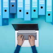 Organized archive with ring binders and woman searching for files in the database using a laptop, top view